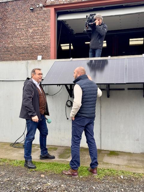 Julien Mawet avec un cameraman et un journaliste de la RTBF, à l'extérieur devant les locaux de Wattuneed, avec des panneaux solaires plug and play en arrière-plan