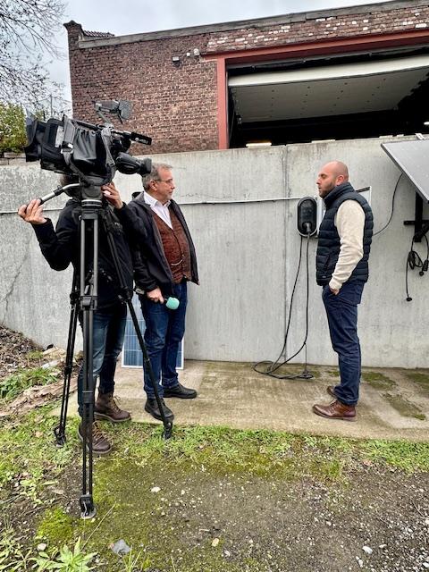 Julien Mawet avec un cameraman et un journaliste de la RTBF, à l'extérieur devant les locaux de Wattuneed, avec des panneaux solaires plug and play en arrière-plan
