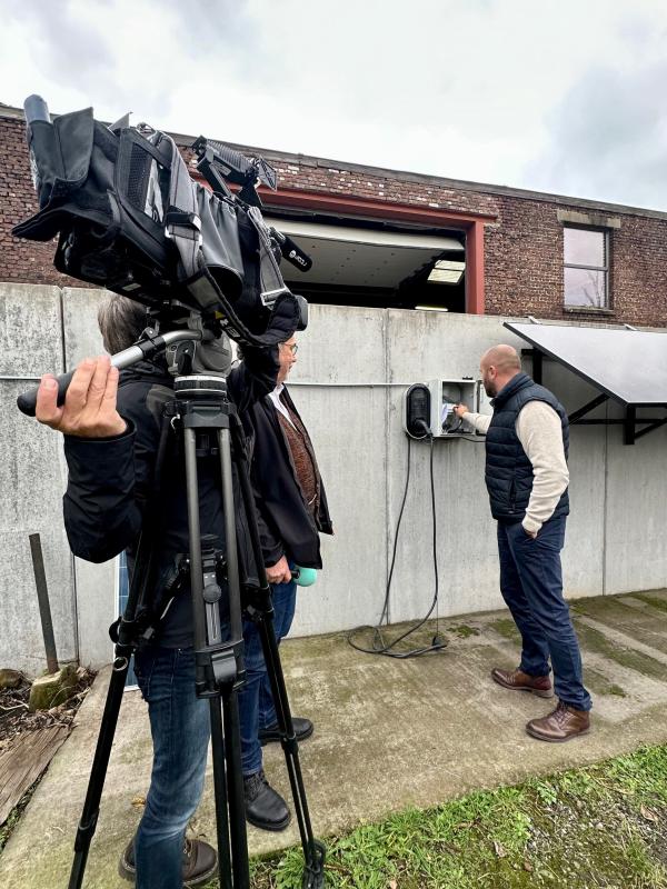Julien Mawet branchant un câble plug and play dans une prise, devant un journaliste et un cameraman, à l'extérieur devant les locaux de Wattuneed, avec des panneaux solaires en arrière-plan