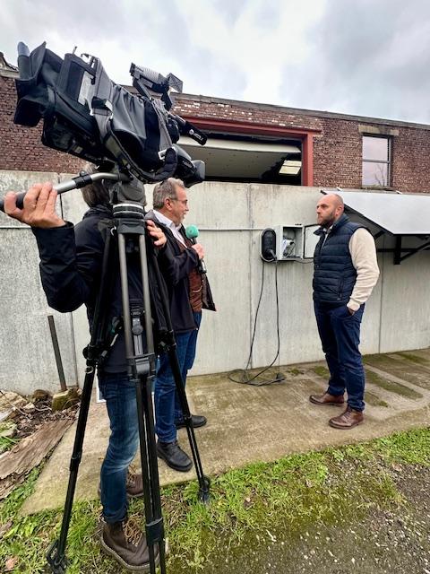 Julien Mawet branchant un câble plug and play dans une prise, devant un journaliste et un cameraman, à l'extérieur devant les locaux de Wattuneed, avec des panneaux solaires en arrière-plan